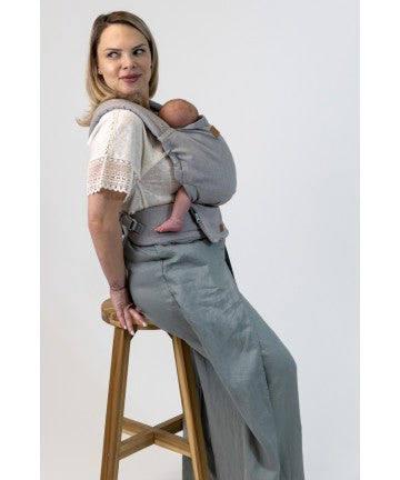 Mulher sentada num banco de madeira a usar a mochila Isara ELITE Misty Linen com um bebé aconchegado, mostrando o conforto e o tecido jacquard.
