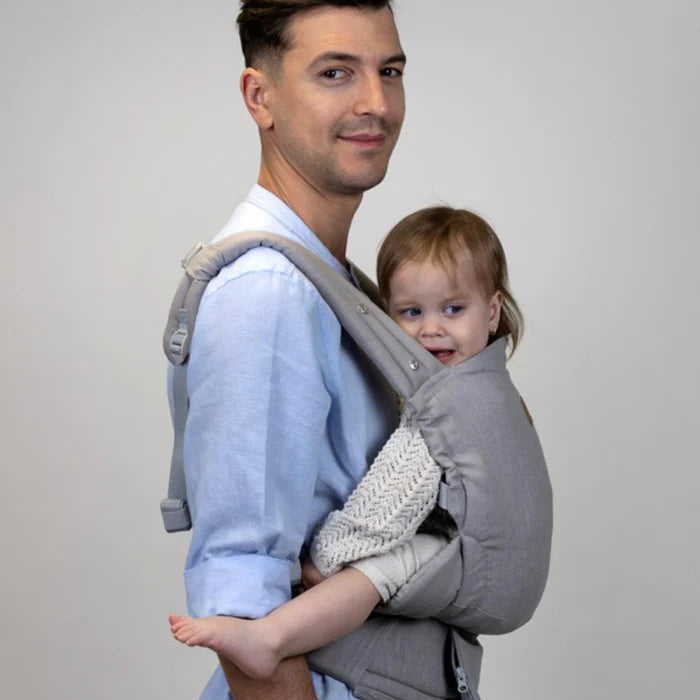 Pai sorridente a usar a mochila Isara ELITE Ajustável Misty Linen com uma criança mais crescida em posição frontal, evidenciando o ajuste para várias idades.