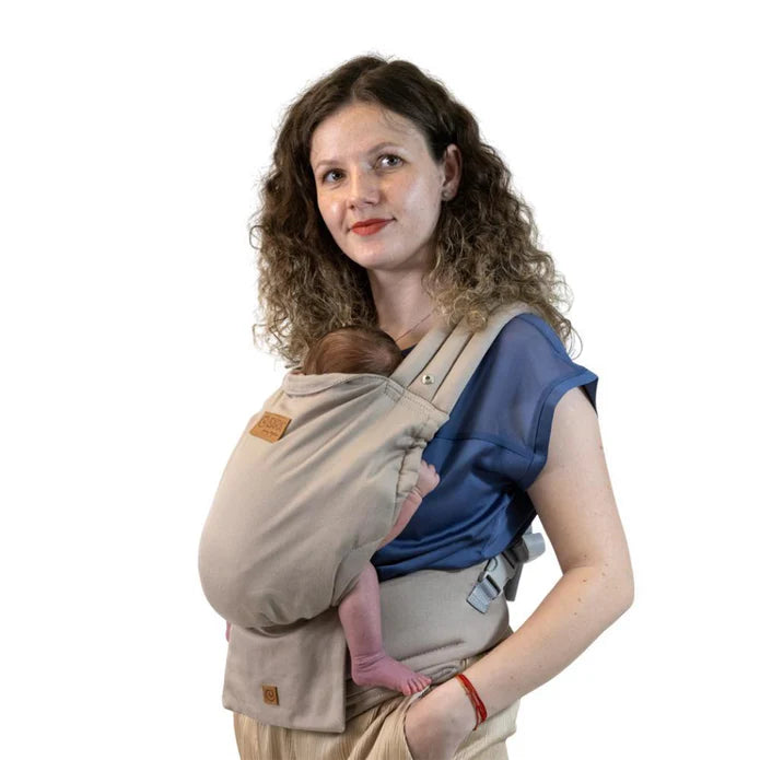 Mulher com cabelo encaracolado a usar uma mochila porta-bebés Isara ELITE bege/castanha clara, transportando um bebé recém-nascido na posição frontal.