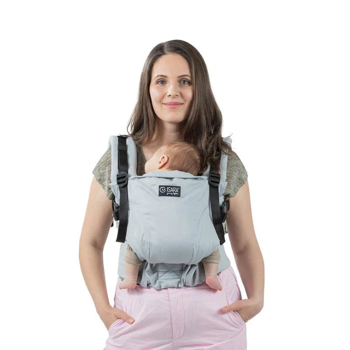 Uma mãe sorridente usando a Mochila Isara THE One ajustável na cor cinzenta pérola, carregando um bebé recém-nascido na posição frontal.
