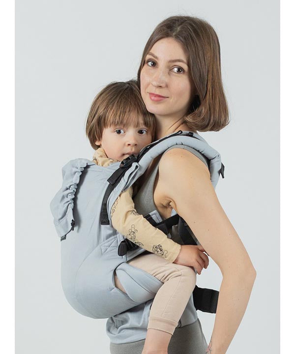 Uma mãe usando a Mochila Isara THE One ajustável na cor cinzenta pérola, carregando uma criança um pouco mais velha na posição nas costas.