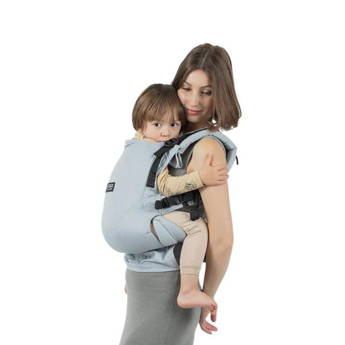 Uma mãe usando a Mochila Isara THE One ajustável na cor cinzenta pérola, carregando uma criança um pouco mais velha na posição lateral.