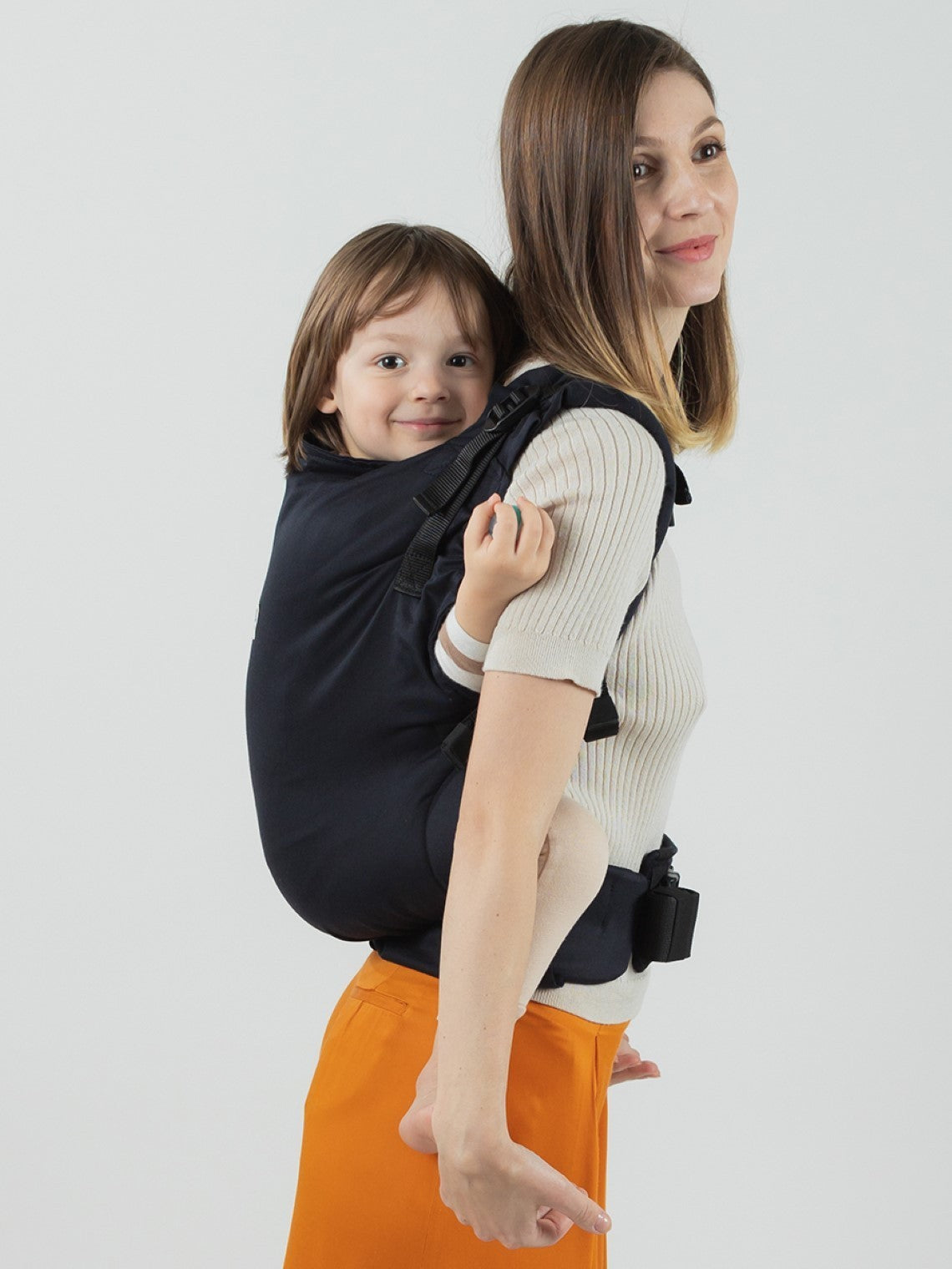 Mulher a usar a mochila Isara Toddler Preschooler Noire com uma criança às costas, vista lateral.