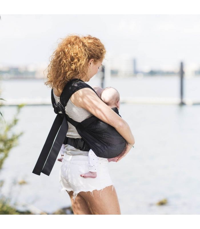 Mulher a carregar dois bebés numa mochila porta-bebés gemelar, ambos posicionados na frente dela, com um sorriso no rosto.