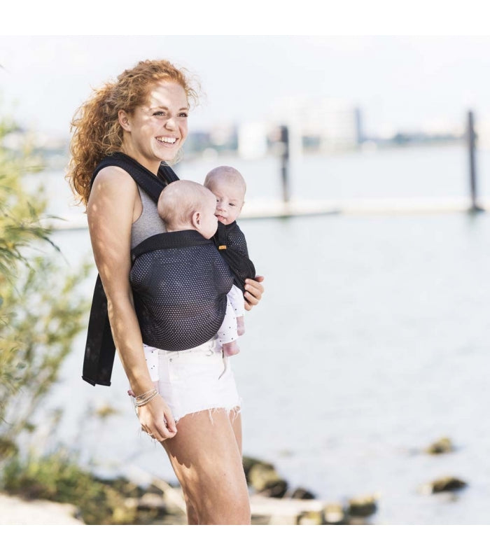 Mulher à beira-mar com a mochila porta-bebés gemelar, dois bebés à frente, numa posição confortável e segura.