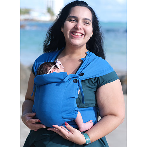 Mulher sorridente transporta um bebé a dormir na mochila Neko Tiny azul vibrante na praia, mostrando o uso exterior.