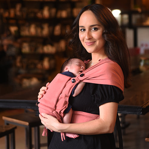 Mãe sorridente transporta um recém-nascido a dormir na mochila Neko Tiny cor de rosa, ajustada ao corpo.