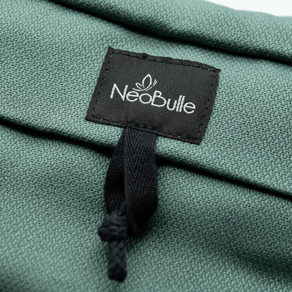 Detalhe macro da etiqueta preta com o logótipo "Neobulle" bordado a branco sobre o tecido texturizado verde da mochila portabebés.