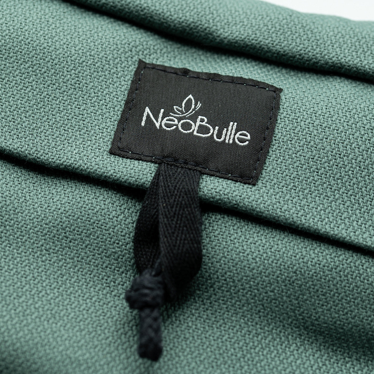 Detalhe macro da etiqueta preta com o logótipo "Neobulle" bordado a branco sobre o tecido texturizado verde da mochila portabebés.