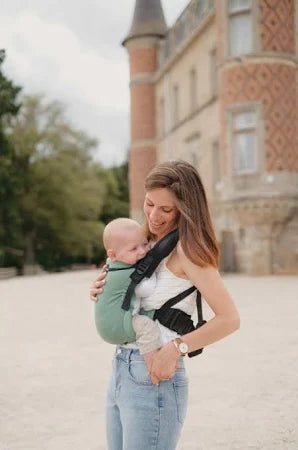 Mãe sorridente a usar a mochila portabebés Neobulle Ajustável ToddlerSize verde, transportando o bebé em frente, com um castelo antigo ao fundo.