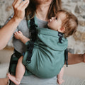 Pai com cabelo comprido a usar a mochila portabebés Neobulle Ajustável ToddlerSize na cor verde, transportando um bebé confortavelmente virado para si.