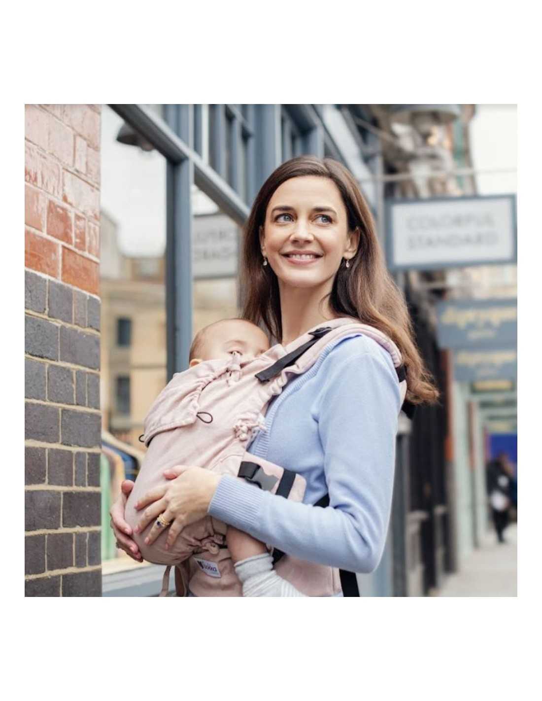 Mulher a utilizar a Mochila Quokkababy E-Carrier Delicious Pink, carregando o seu bebé na posição frontal enquanto sorri. A imagem destaca a praticidade e conforto da mochila em uso diário.