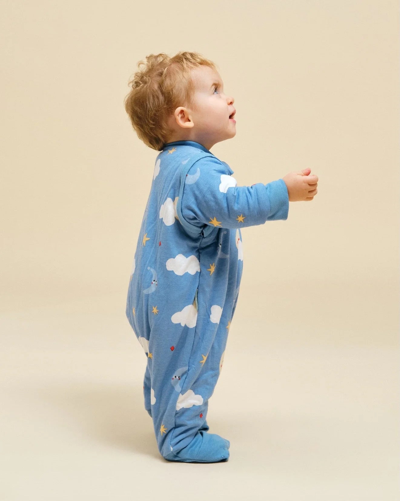 Bebé de pé, de perfil, a olhar para cima, vestindo o saco de dormir MolisandCo TOG 1.0 azul com mangas e pés, padrão "Good Night".