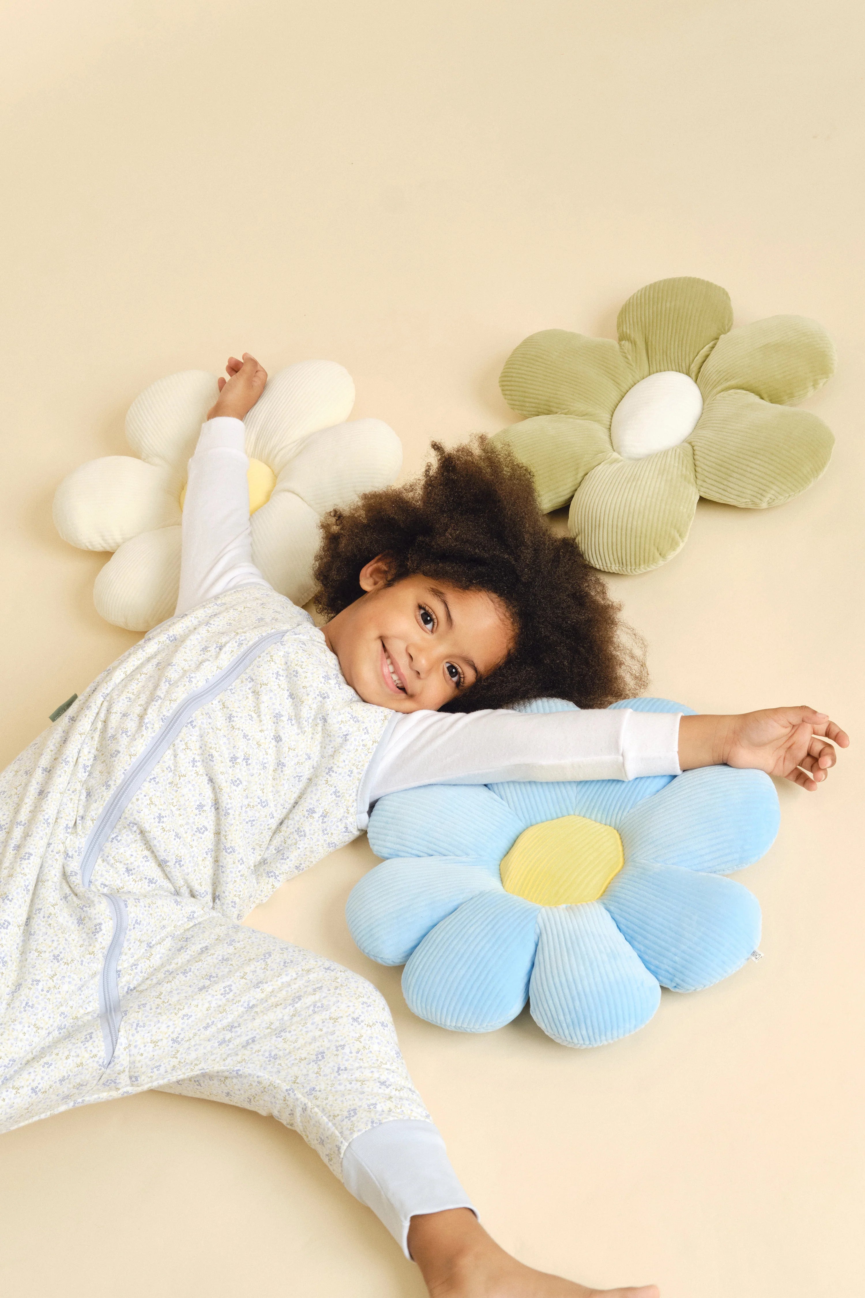 Criança sorridente deitada no chão com o saco de dormir MolisandCo, rodeada por almofadas decorativas em forma de flor.
