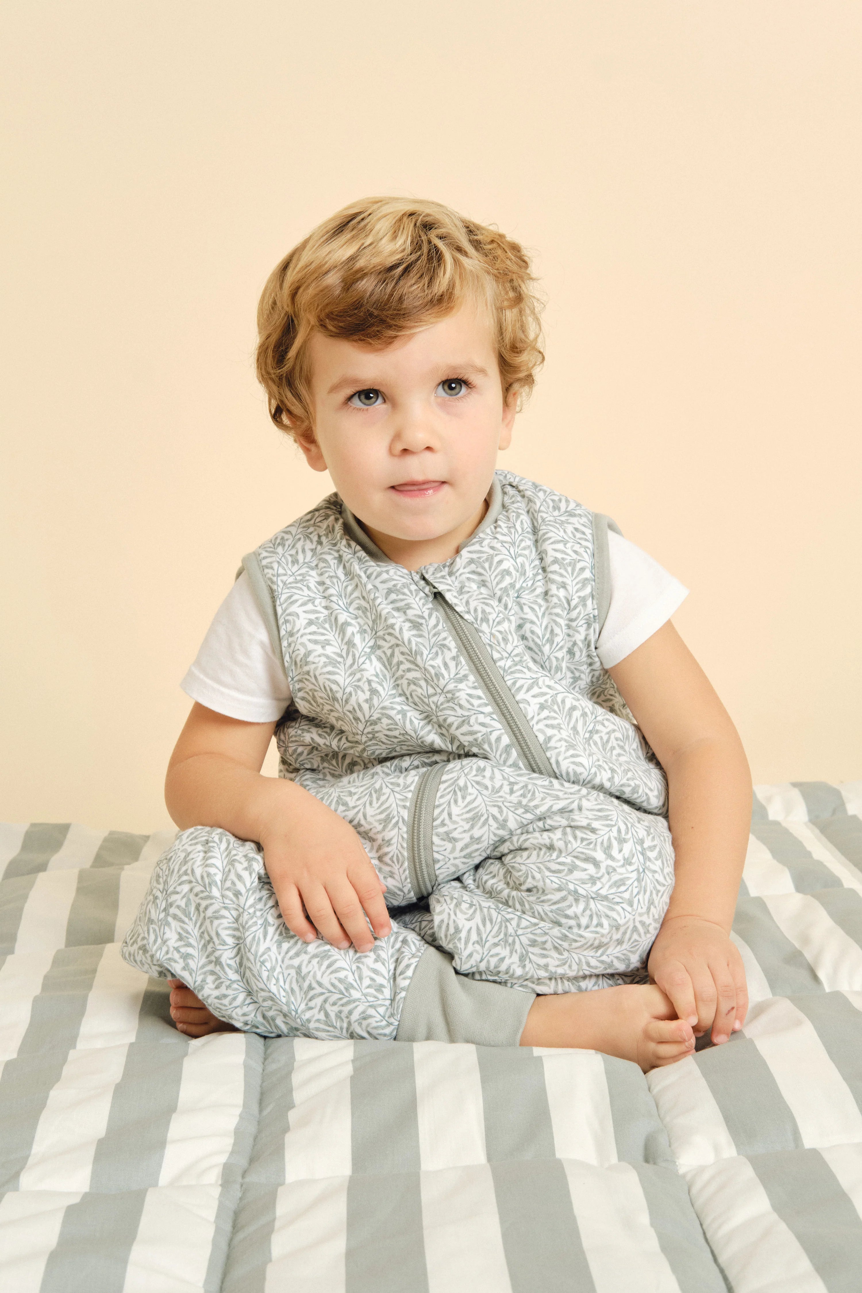 Criança de cabelo loiro e olhos azuis, vestindo o saco de dormir com pés MolisandCo INES II, sentada sobre um tapete listrado a brincar.