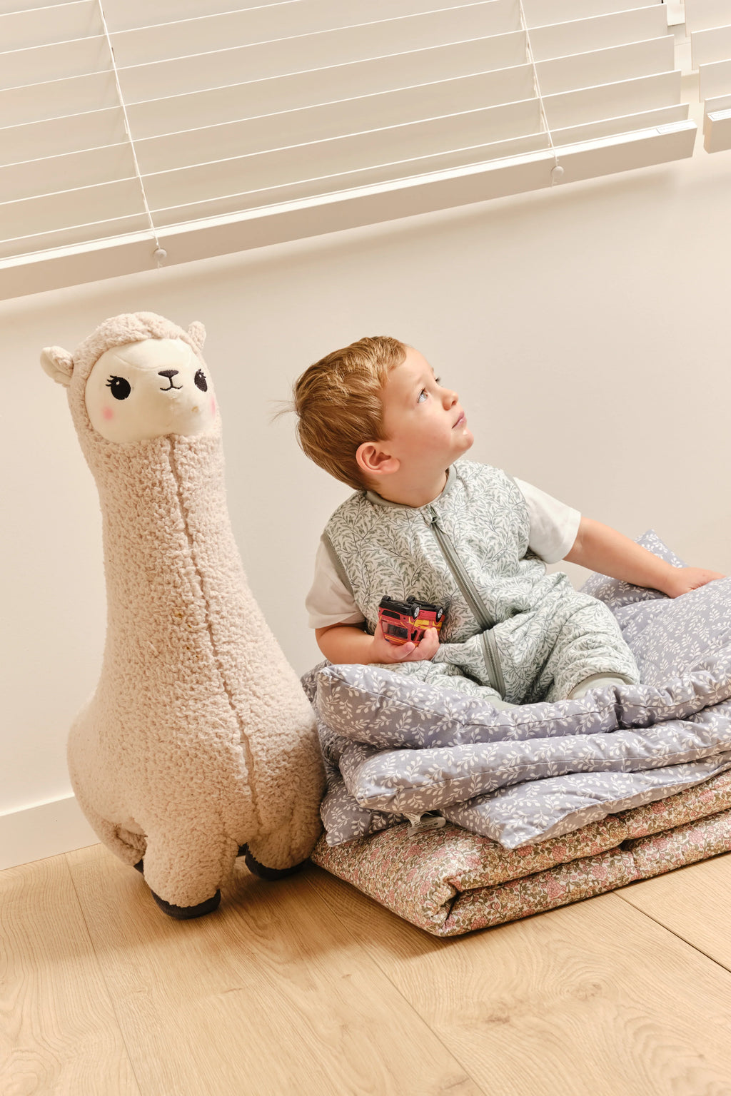 Criança no saco de dormir MolisandCo INES II ao lado de um peluche de lhama, sentada sobre pilhas de cobertores florais e lisos.