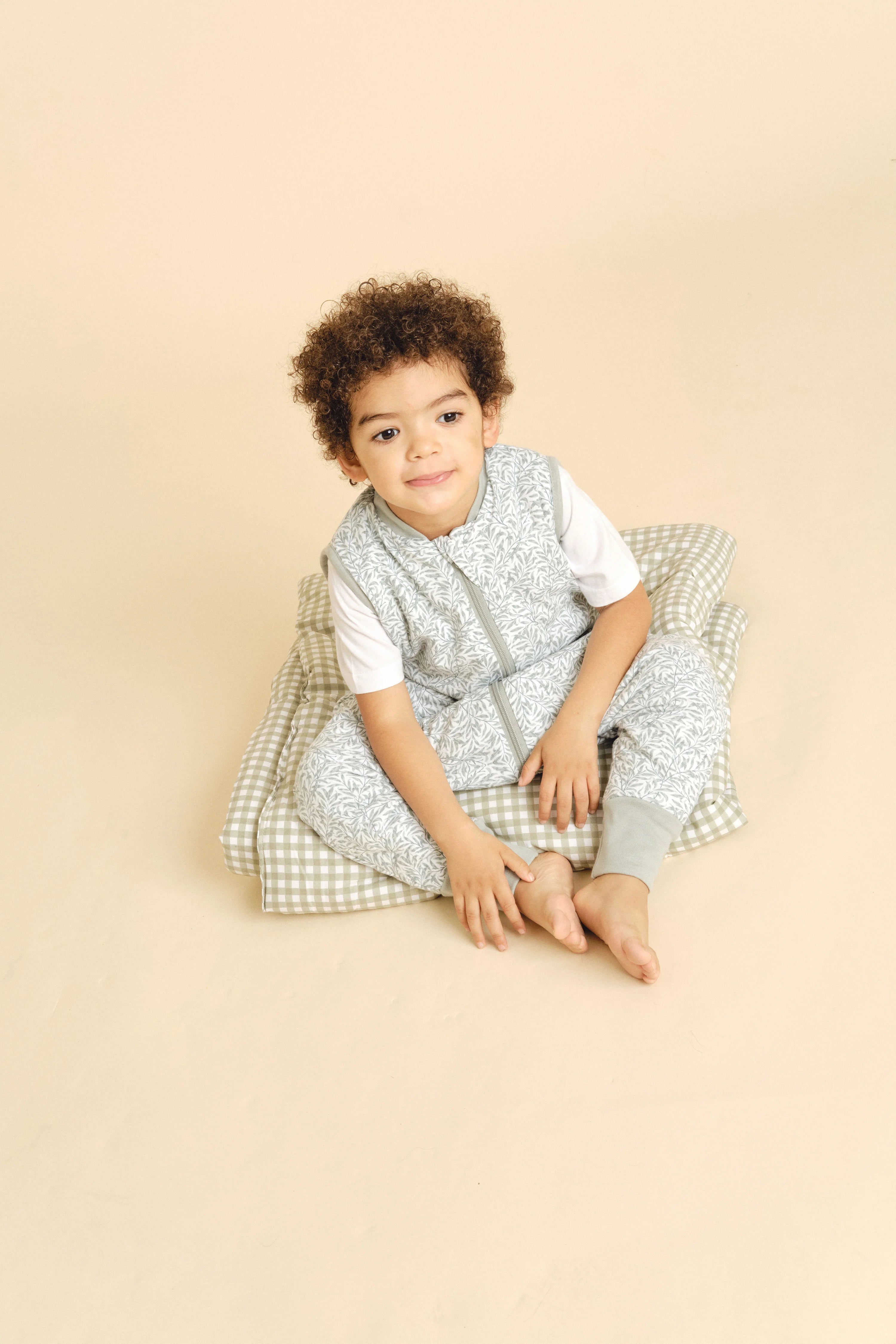 Menino com cabelo encaracolado a usar o saco de dormir com pés MolisandCo INES II, sentado confortavelmente numa almofada xadrez verde-clara.