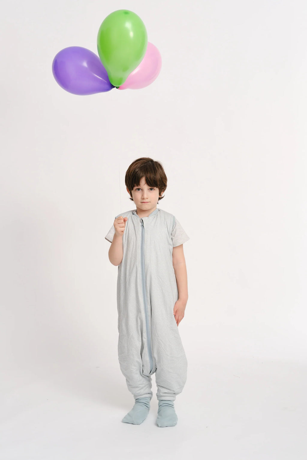 Criança em pé, vestindo o saco de dormir MolisandCo TOG 1.0 com pés, a segurar um balão roxo, com balões verde e rosa ao fundo.