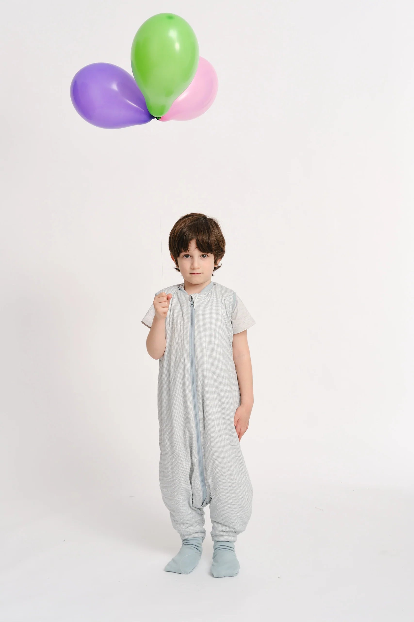 Criança em pé, vestindo o saco de dormir MolisandCo TOG 1.0 com pés, a segurar um balão roxo, com balões verde e rosa ao fundo.