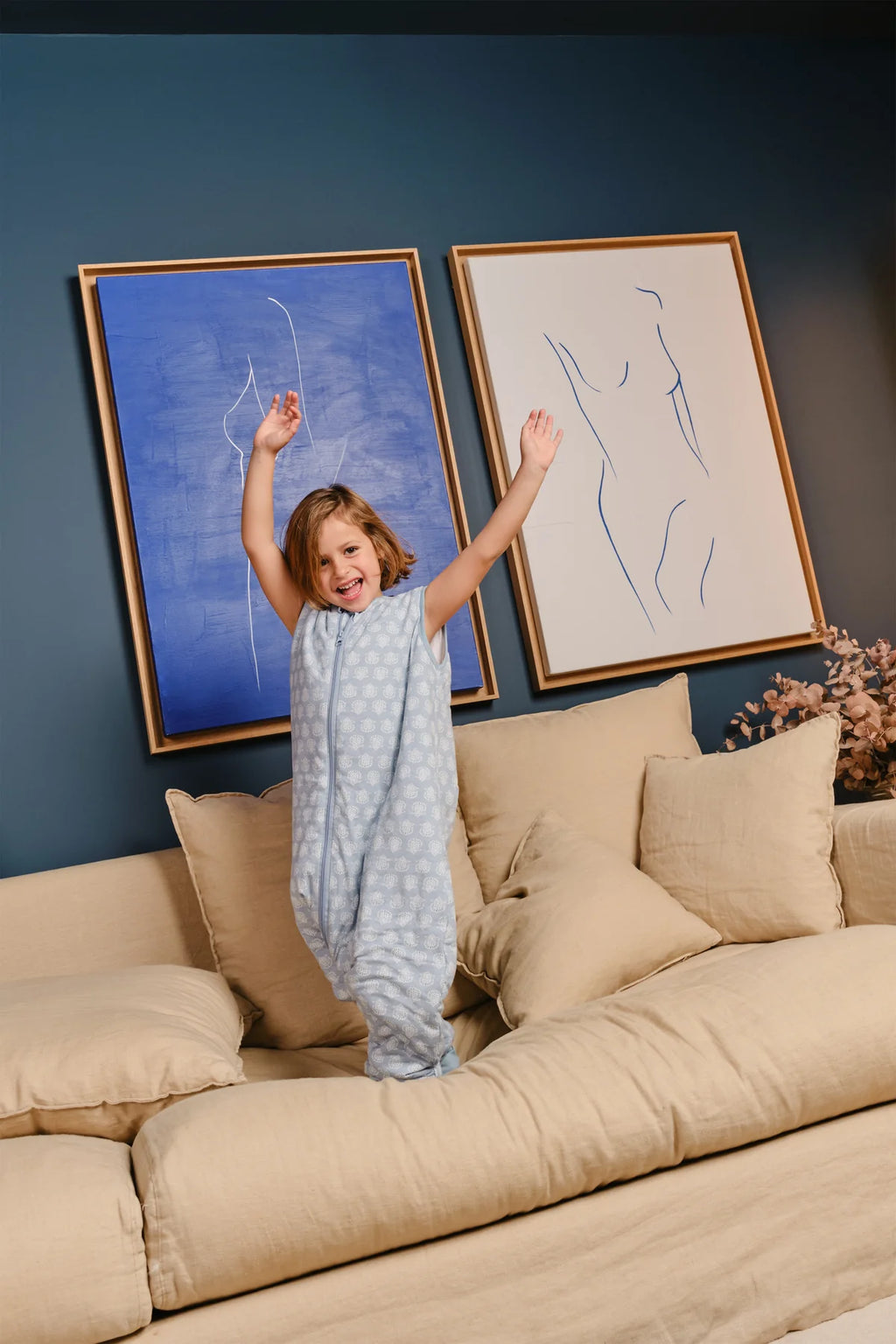 Criança sorridente em pé sobre um sofá bege, vestindo o saco de dormir com pés MolisandCo TOG 1.0 azul claro estampado.