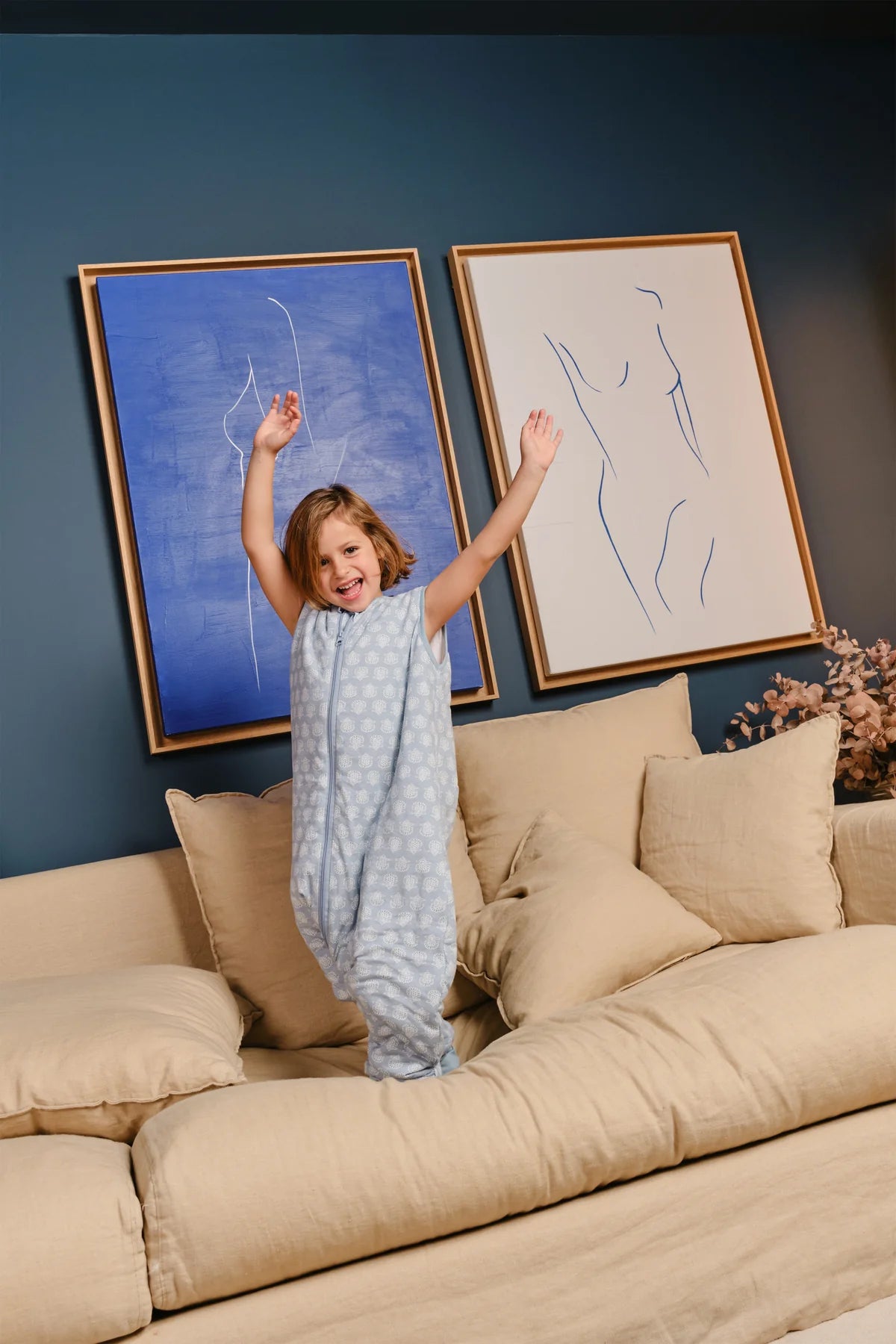 Criança sorridente em pé sobre um sofá bege, vestindo o saco de dormir com pés MolisandCo TOG 1.0 azul claro estampado.