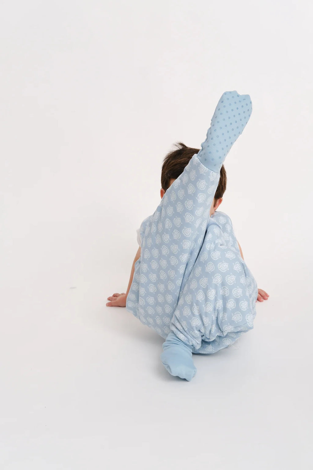 Criança de costas, sentada no chão branco, a levantar uma perna coberta pelo saco de dormir com pés azul claro estampado, mostrando a sola antiderrapante.