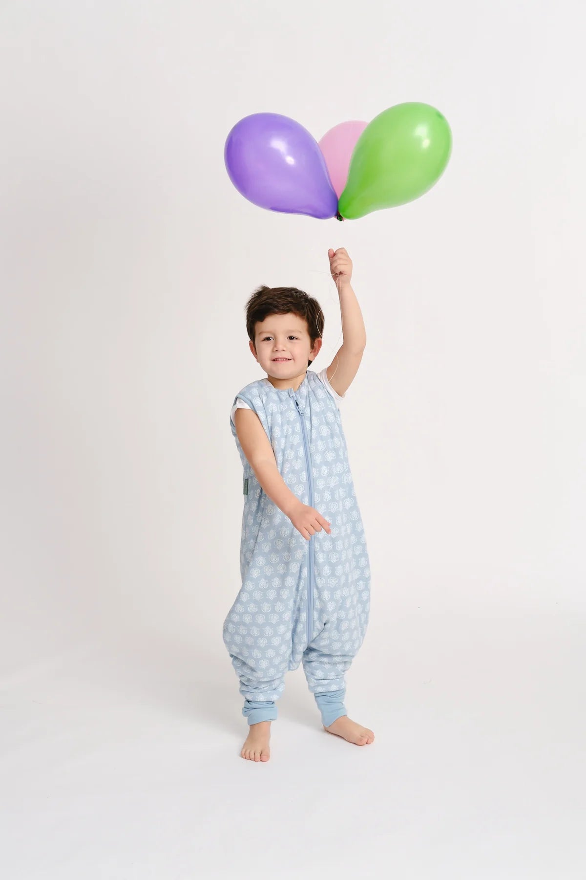 Menino sorridente em estúdio, vestido com o saco de dormir com pés MolisandCo TOG 1.0 azul claro, a segurar três balões coloridos (roxo, rosa e verde).