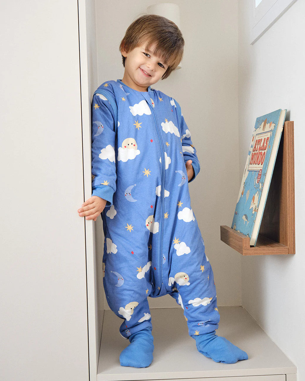 Criança sorridente em pé num nicho, vestindo o saco de dormir MolisandCo TOG 1.0 azul com mangas e pés, padrão "Good Night".