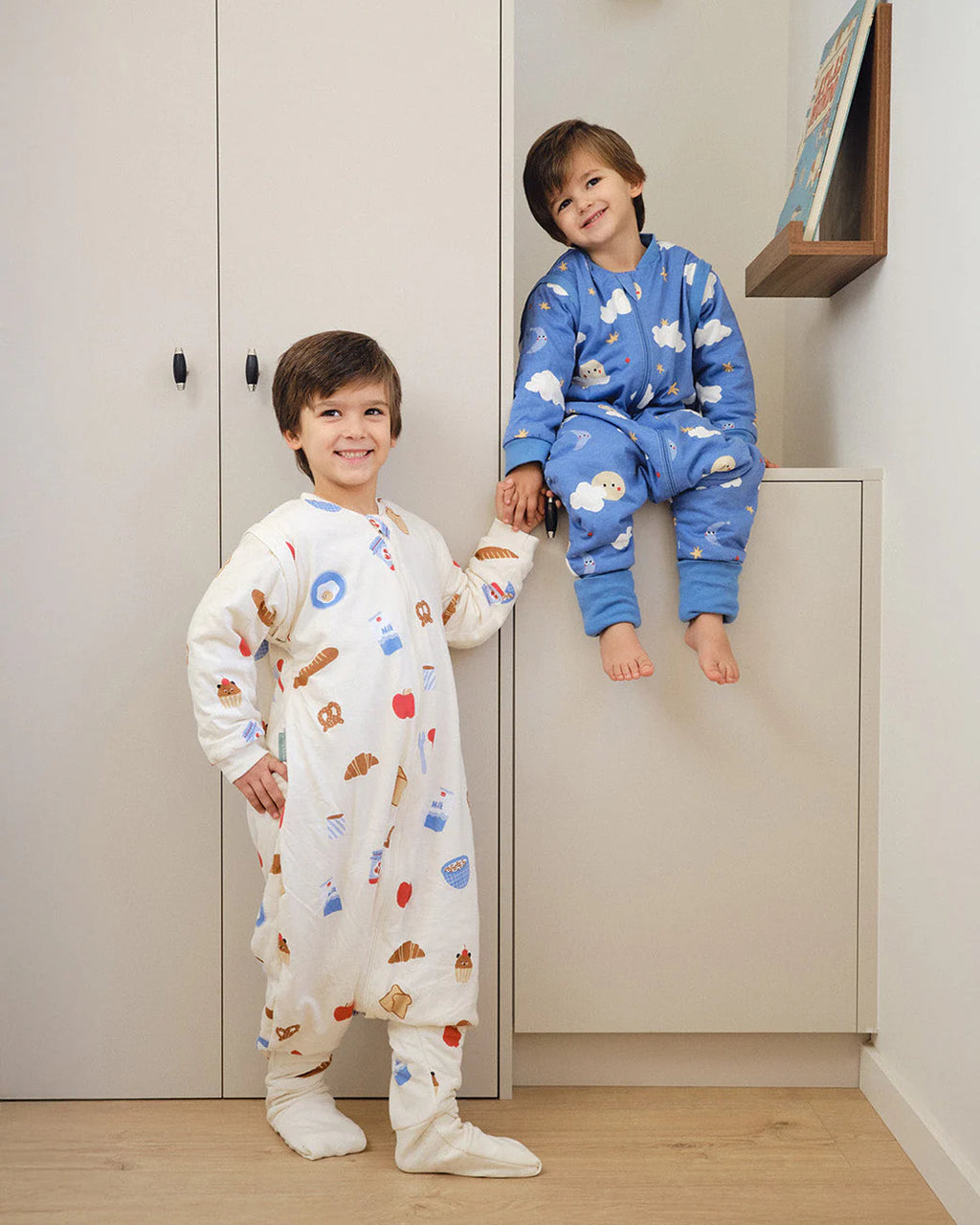 Dois irmãos sorridentes em casa, um de pé com saco de dormir creme (outra estampa) e outro sentado com o saco de dormir azul "Good Night".