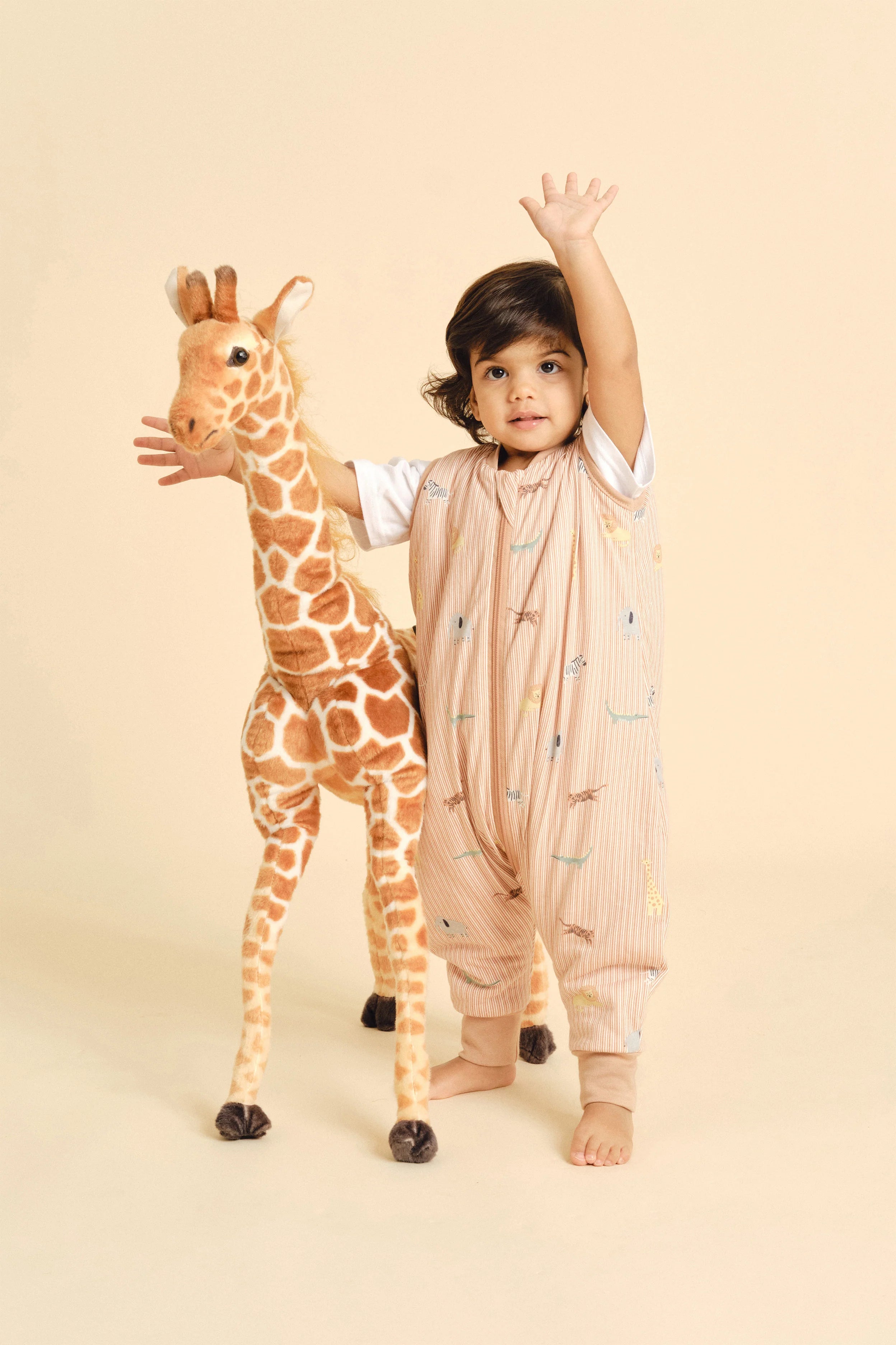Bebé em pé com saco de dormir MolisandCo TOG 1.0 Jungle, braço levantado, ao lado de girafa de peluche.