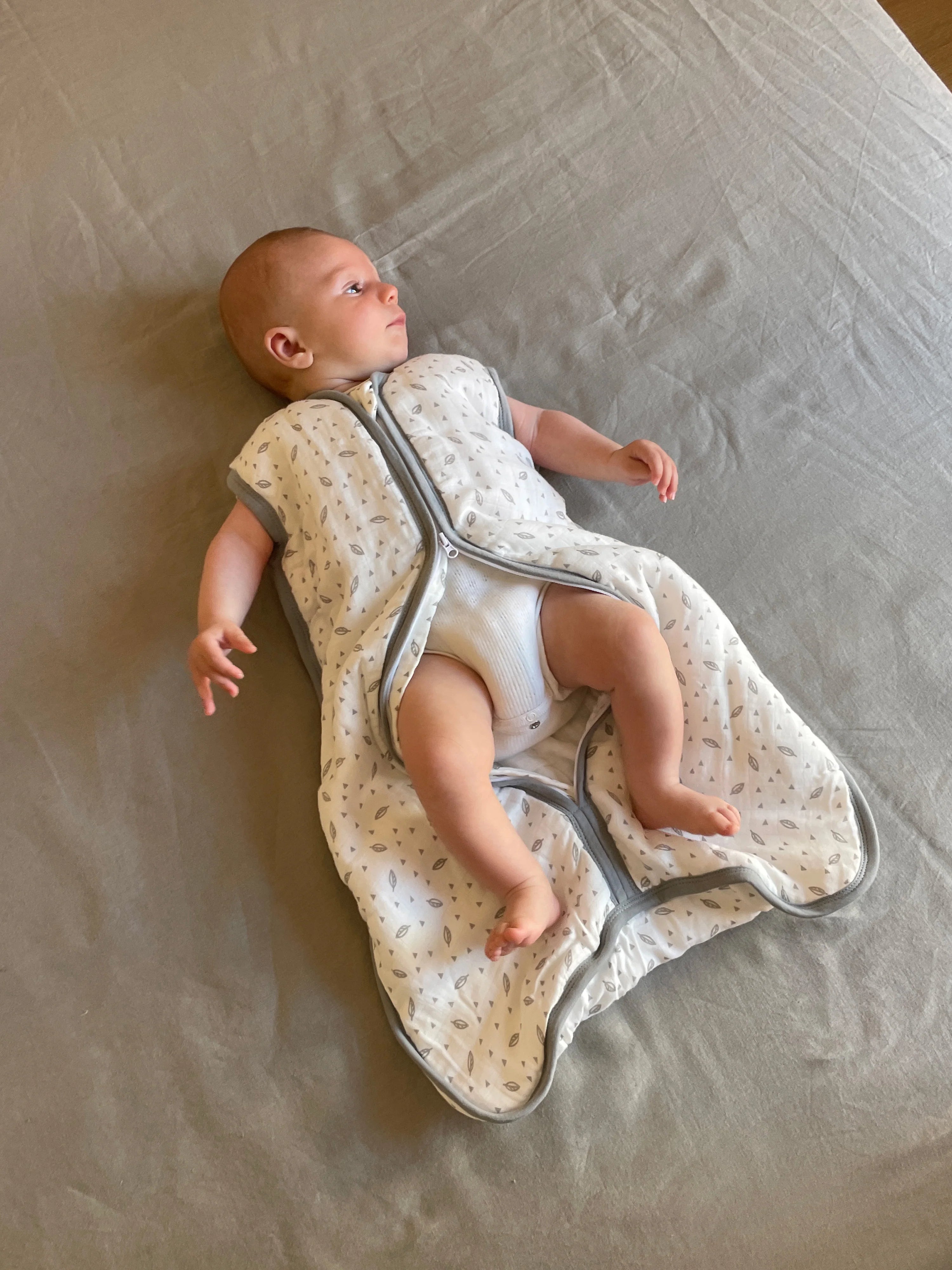 Bebé deitado em lençol cinzento, a usar saco de dormir MolisandCo TOG 1.0 aberto, mostrando pernas e fralda.