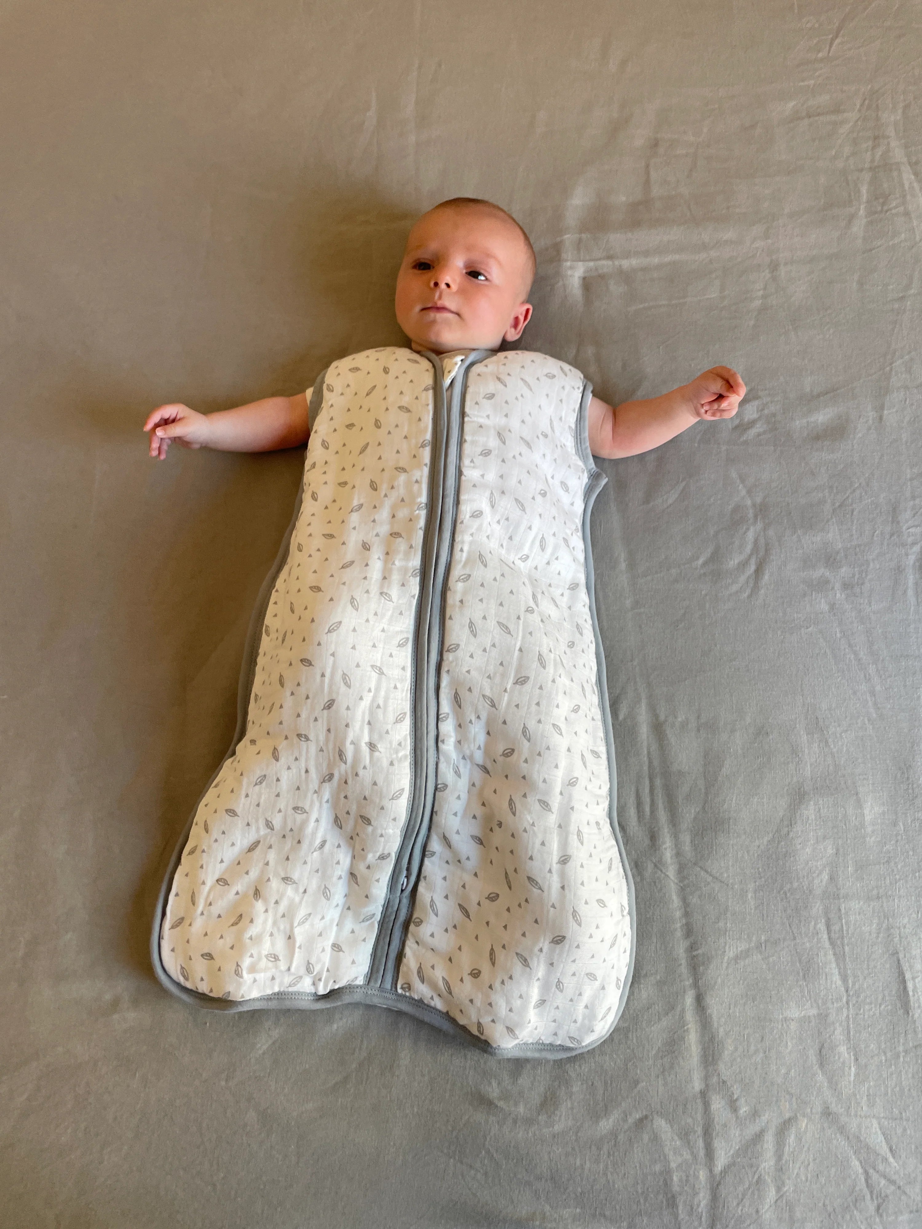 Bebé deitado de costas, a usar saco de dormir MolisandCo TOG 1.0 de musselina com padrão de folhas, fechado.