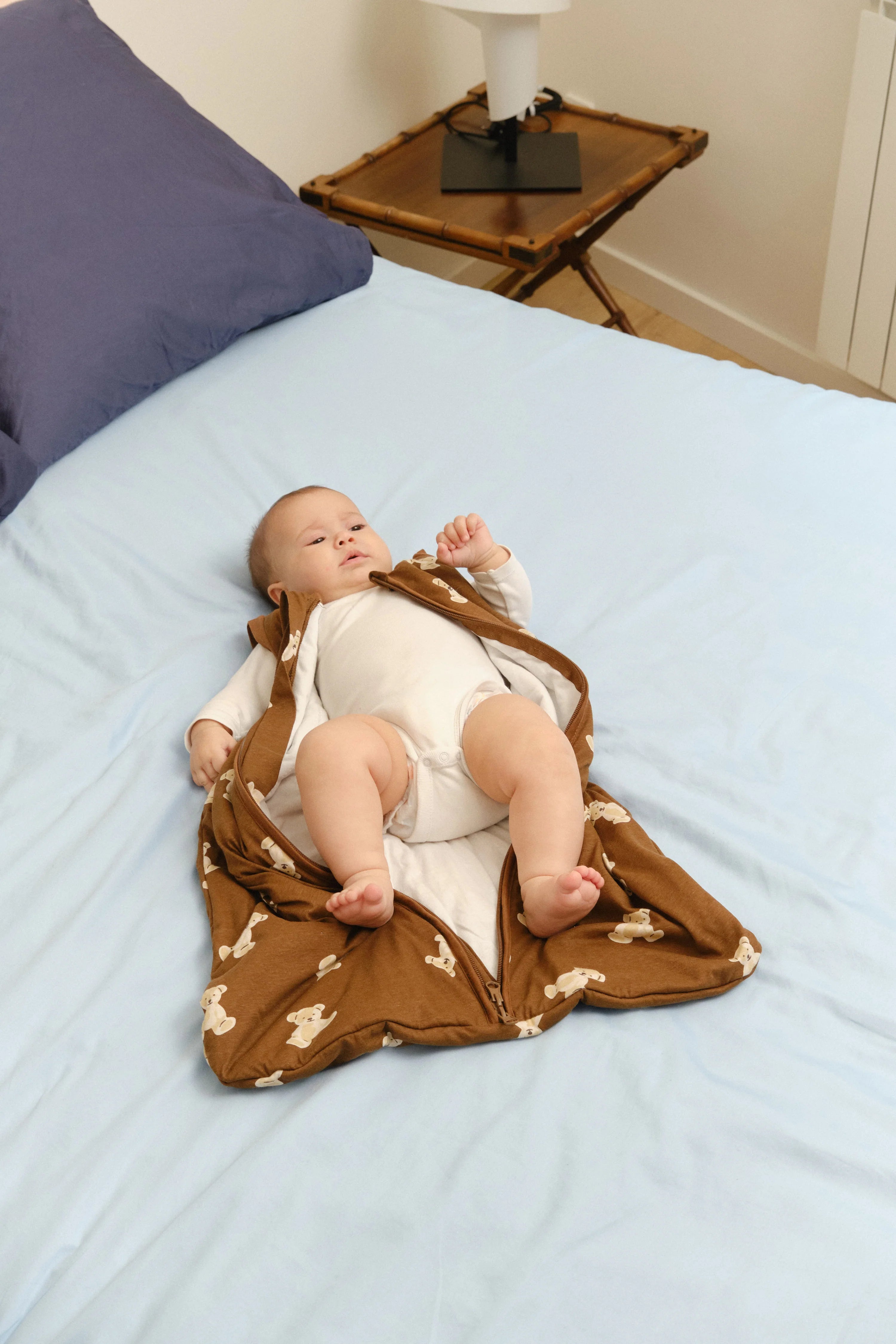 Um bebé deitado de costas numa cama com lençóis azuis, com o saco de dormir MolisandCo TOG 1.0 Teddy Mocca castanho aberto, mostrando um body branco por baixo.