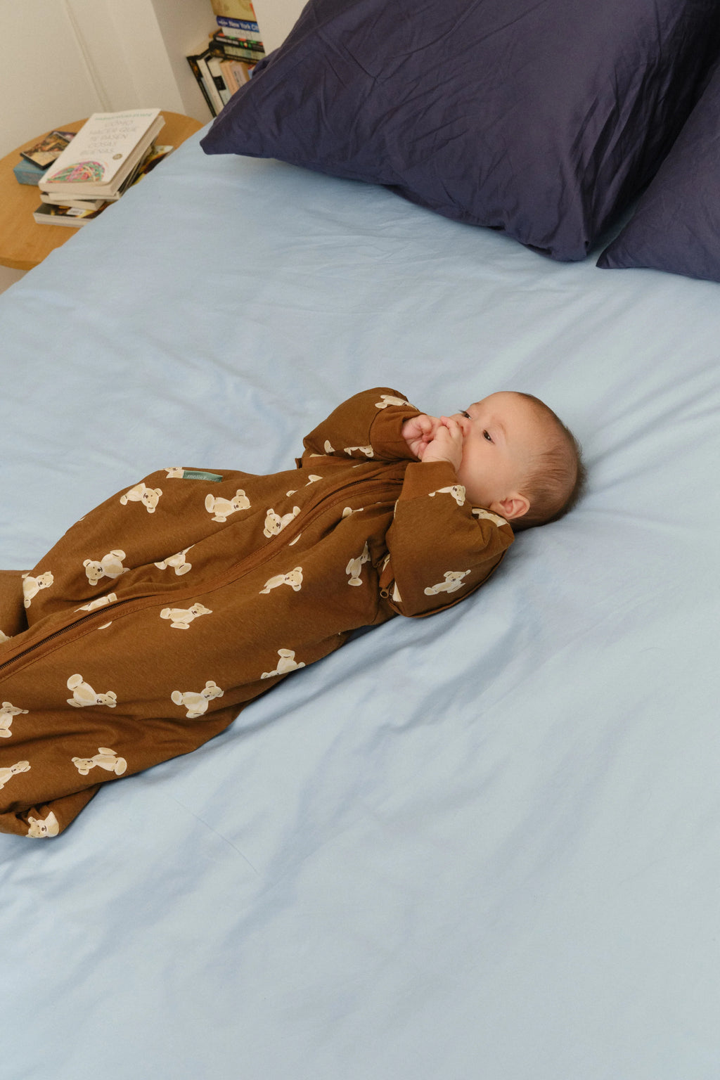 Um bebé deitado de costas numa cama com lençóis azuis, usando o saco de dormir MolisandCo TOG 1.0 Teddy Mocca castanho, com as mãos na boca.