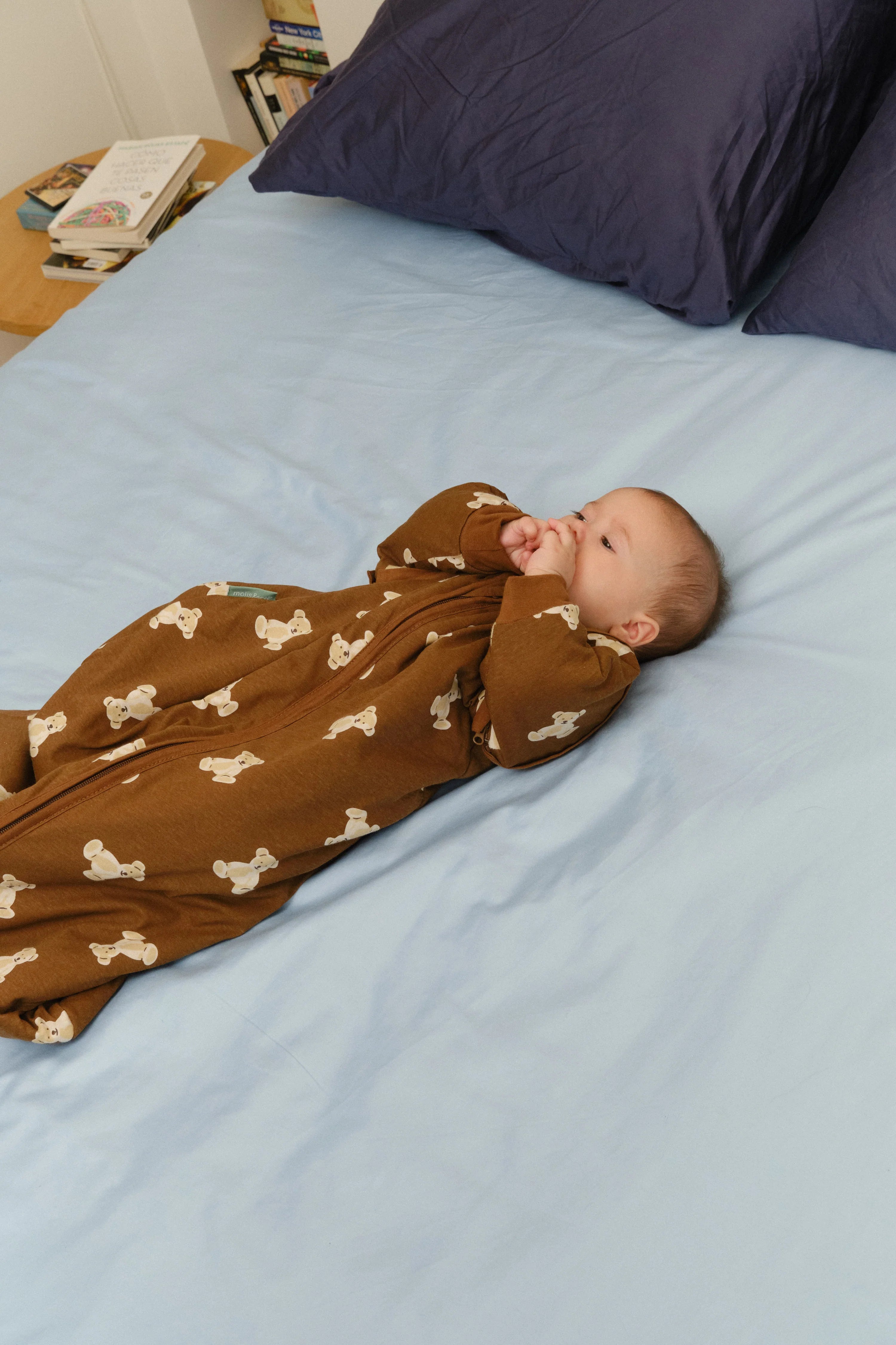 Um bebé deitado de costas numa cama com lençóis azuis, usando o saco de dormir MolisandCo TOG 1.0 Teddy Mocca castanho, com as mãos na boca.