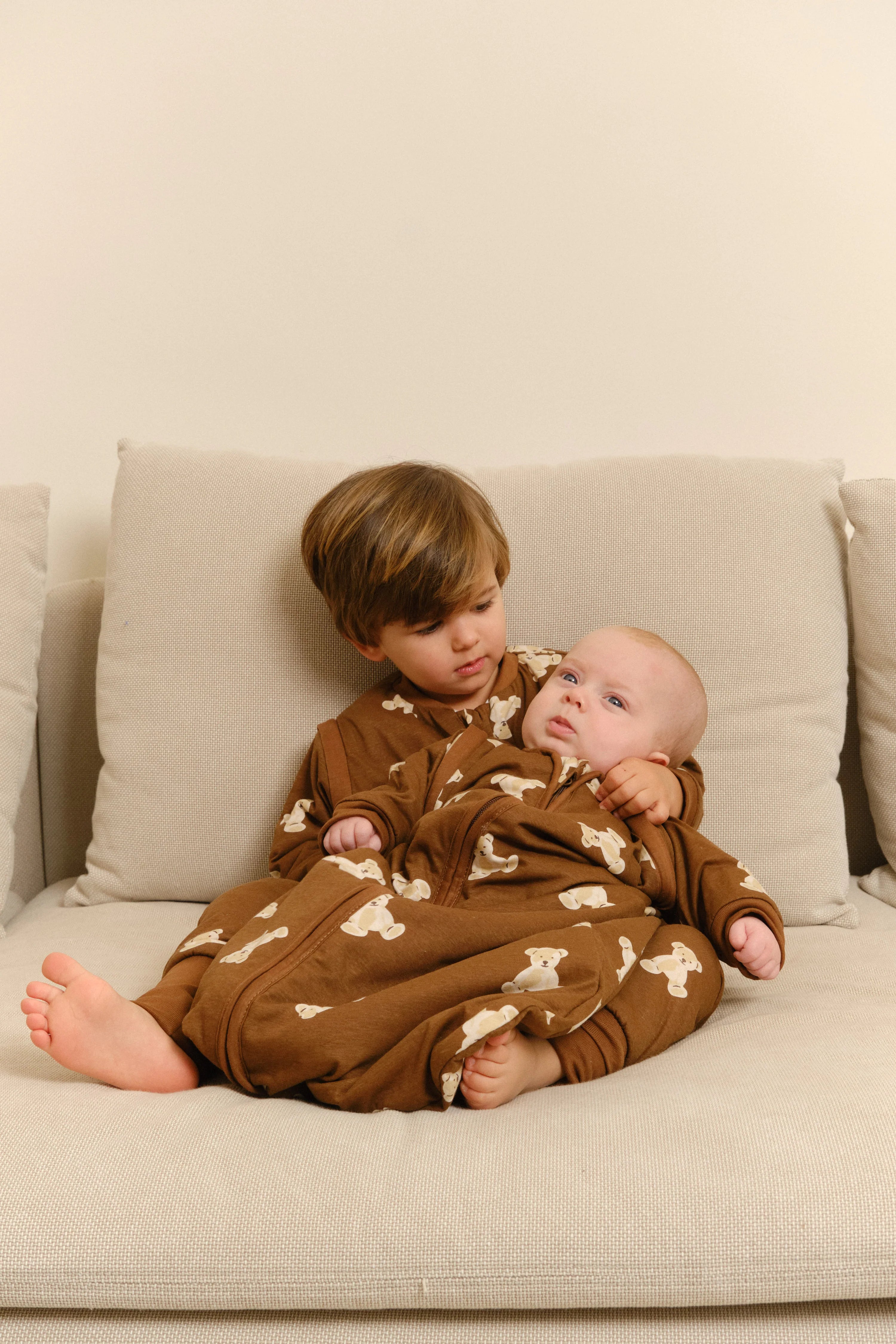 Uma criança mais velha abraça um bebé, ambos vestidos com o saco de dormir MolisandCo TOG 1.0 Teddy Mocca castanho, sentados num sofá.