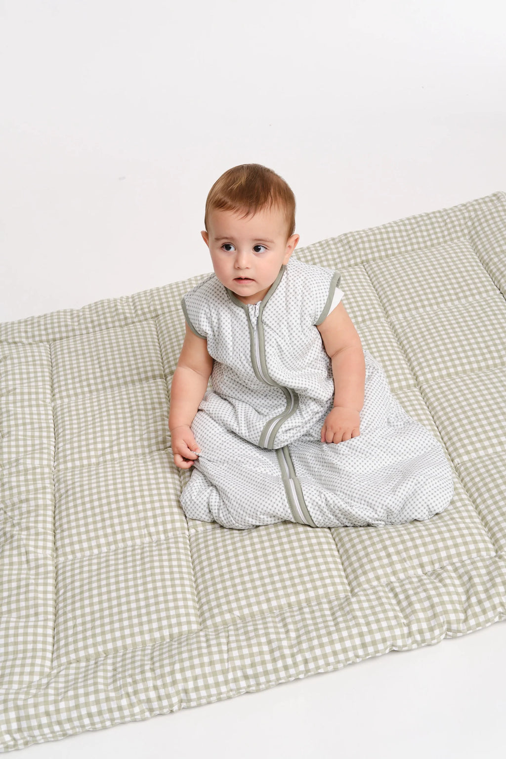 Bebé sentado sobre um tapete xadrez verde e branco, vestindo o saco de dormir MolisandCo TOG 1.0 sem pés, fundo branco.