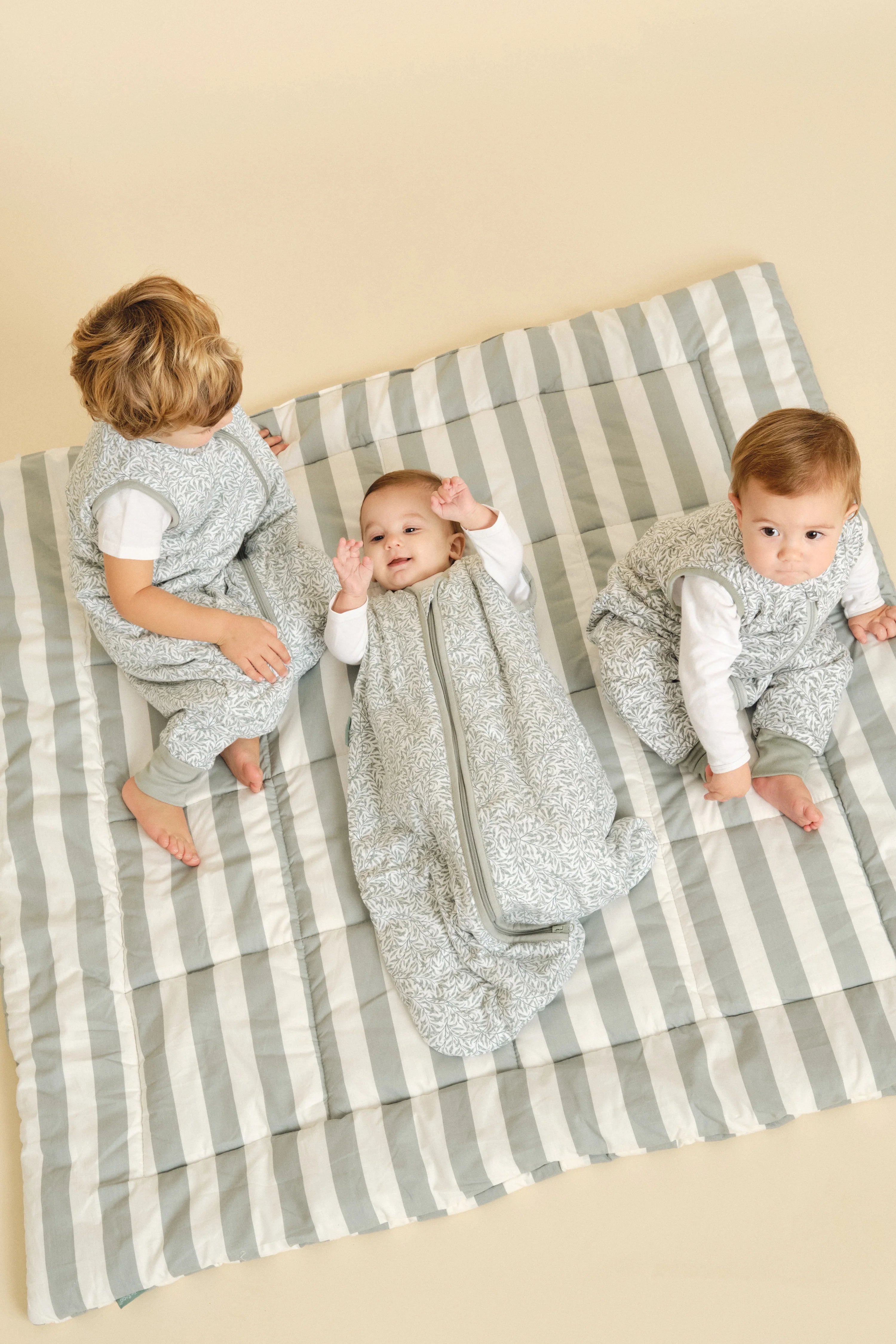 Três bebés em sacos de dormir MolisandCo TOG 1.0 Ines II, um deitado e dois sentados, sobre uma manta às riscas cinzentas.