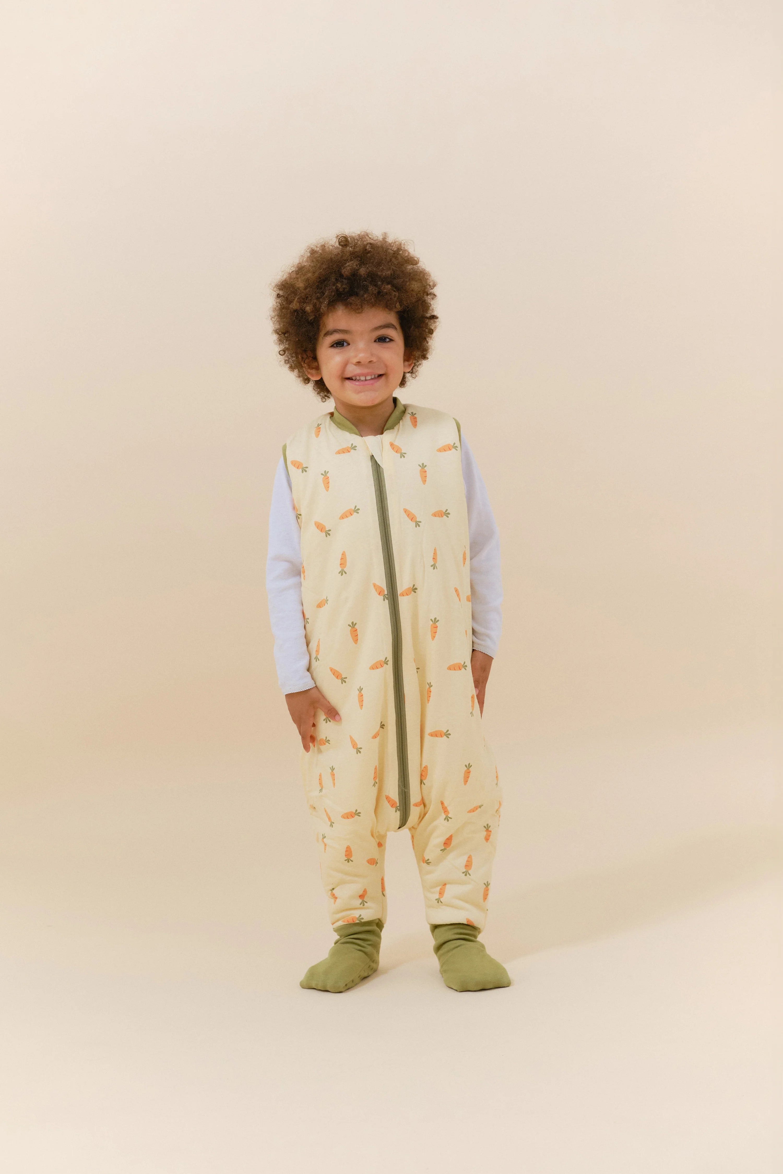 Bebé a sorrir, de pé, com saco de dormir MolisandCo Carrots TOG 2.5 e meias antiderrapantes.