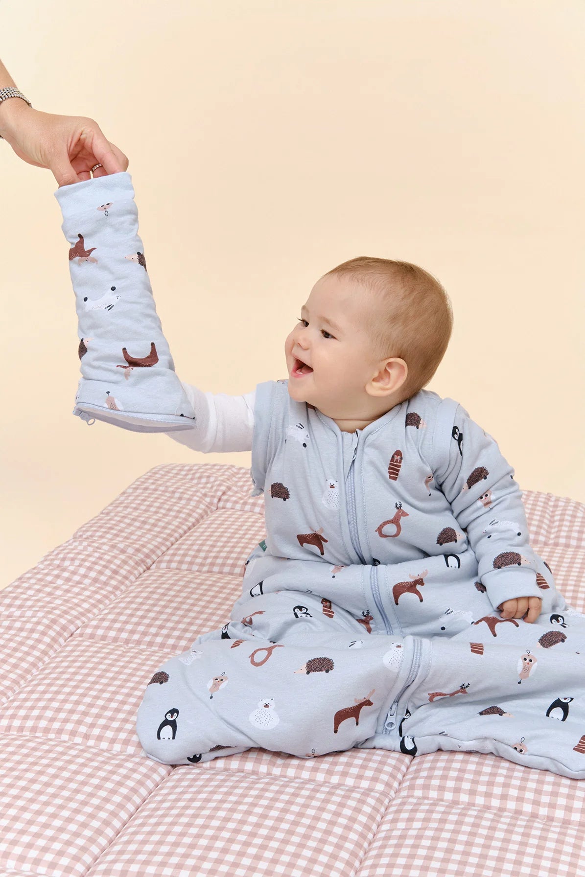 Bebé sorridente sentado, a interagir com uma mão adulta que lhe mostra uma das mangas removíveis do saco de dormir TOG 2.5 "Winter Animals".