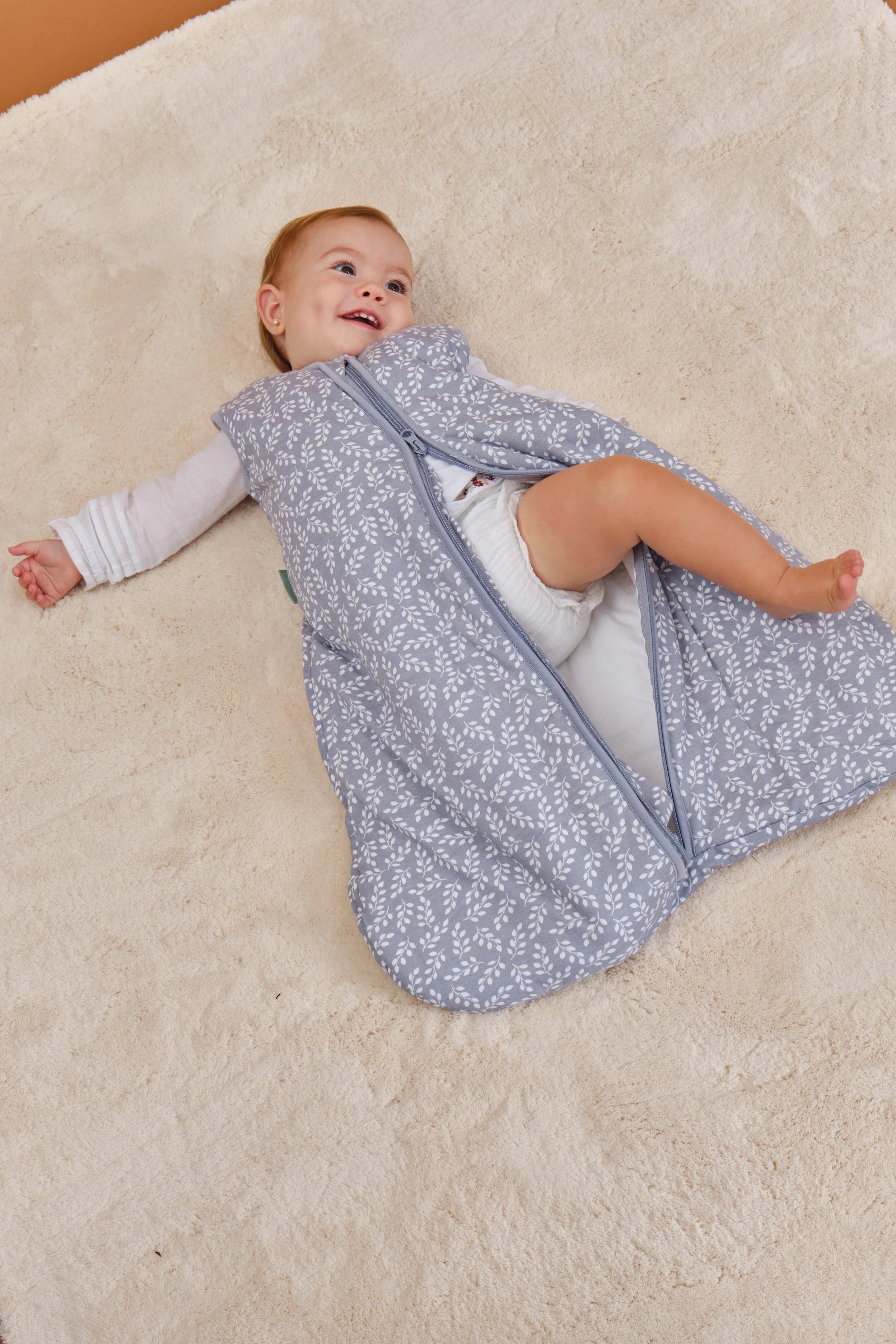 Bebé feliz no chão com saco de dormir MolisandCo TOG 2.5 Blue Garden aberto, mostrando a facilidade de uso.