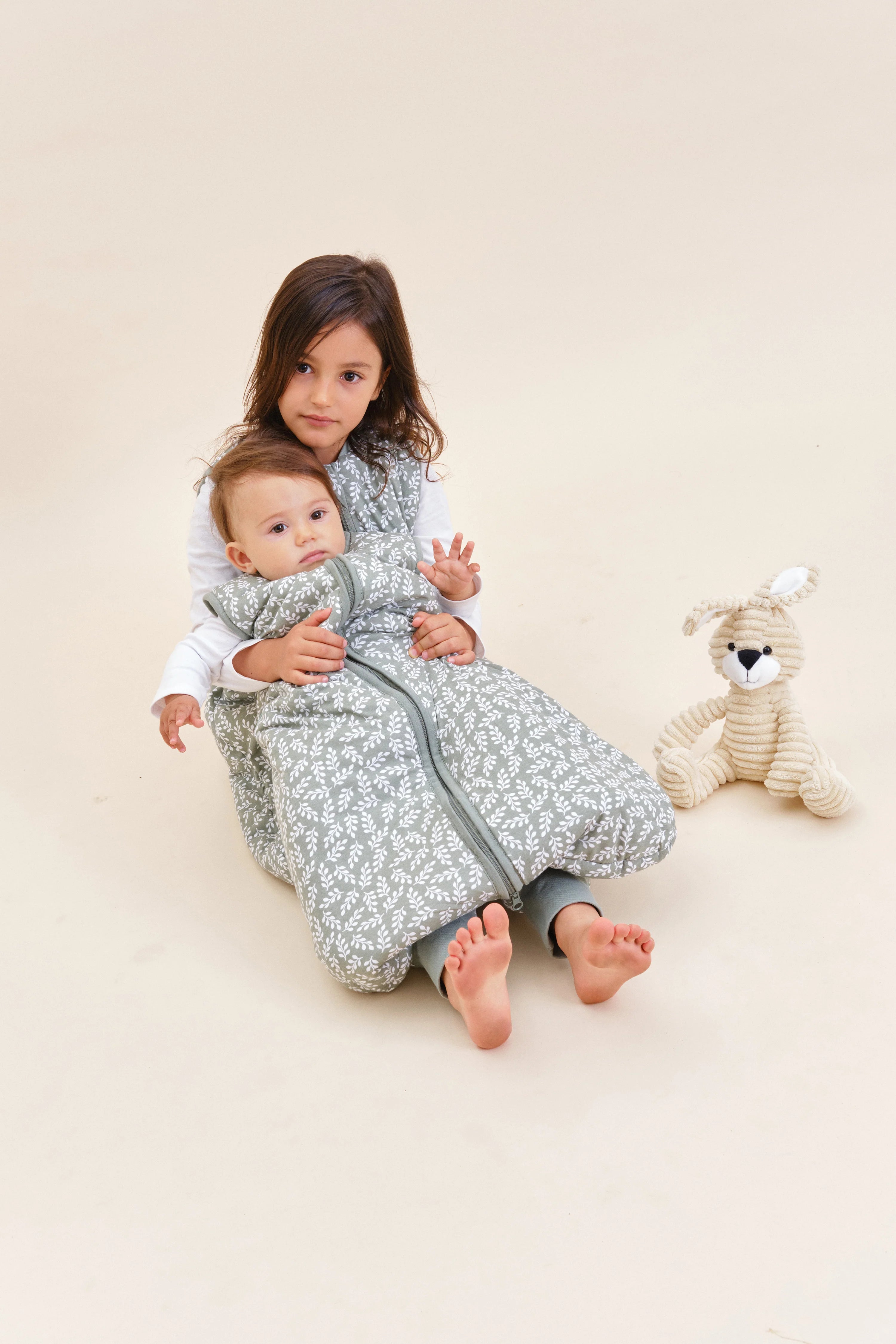 Criança abraça bebé no saco de dormir MolisandCo TOG 2.5 Green Garden, com coelho de peluche ao lado.