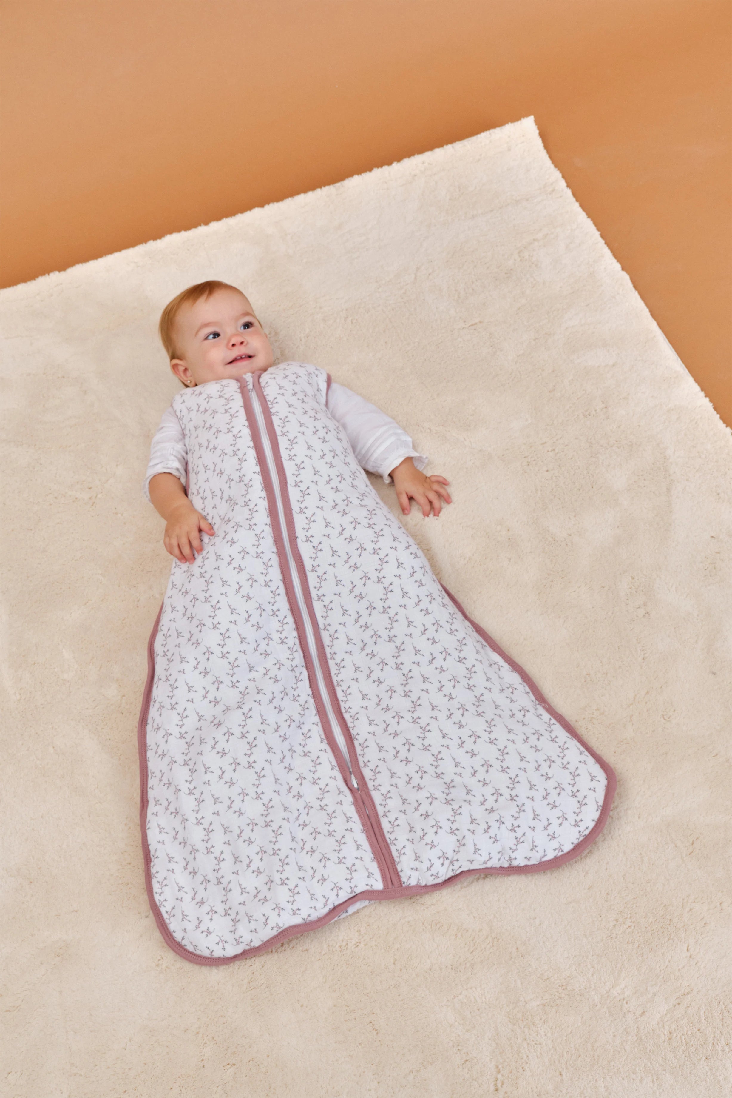 Bebé sorridente deitado no tapete, a usar o saco de dormir MolisandCo TOG 2.5 rosa floral.