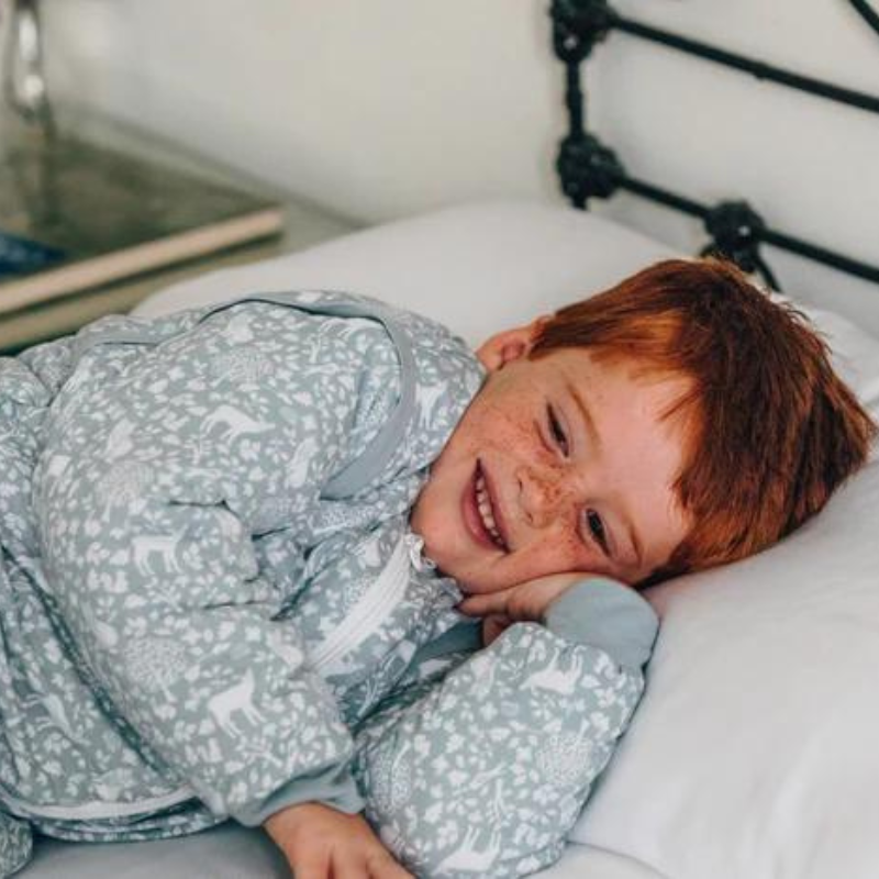 Criança sorridente com cabelo ruivo deitada confortavelmente na cama, vestindo o saco de dormir MolisandCo TOG 2.5 estampado Woodland.
