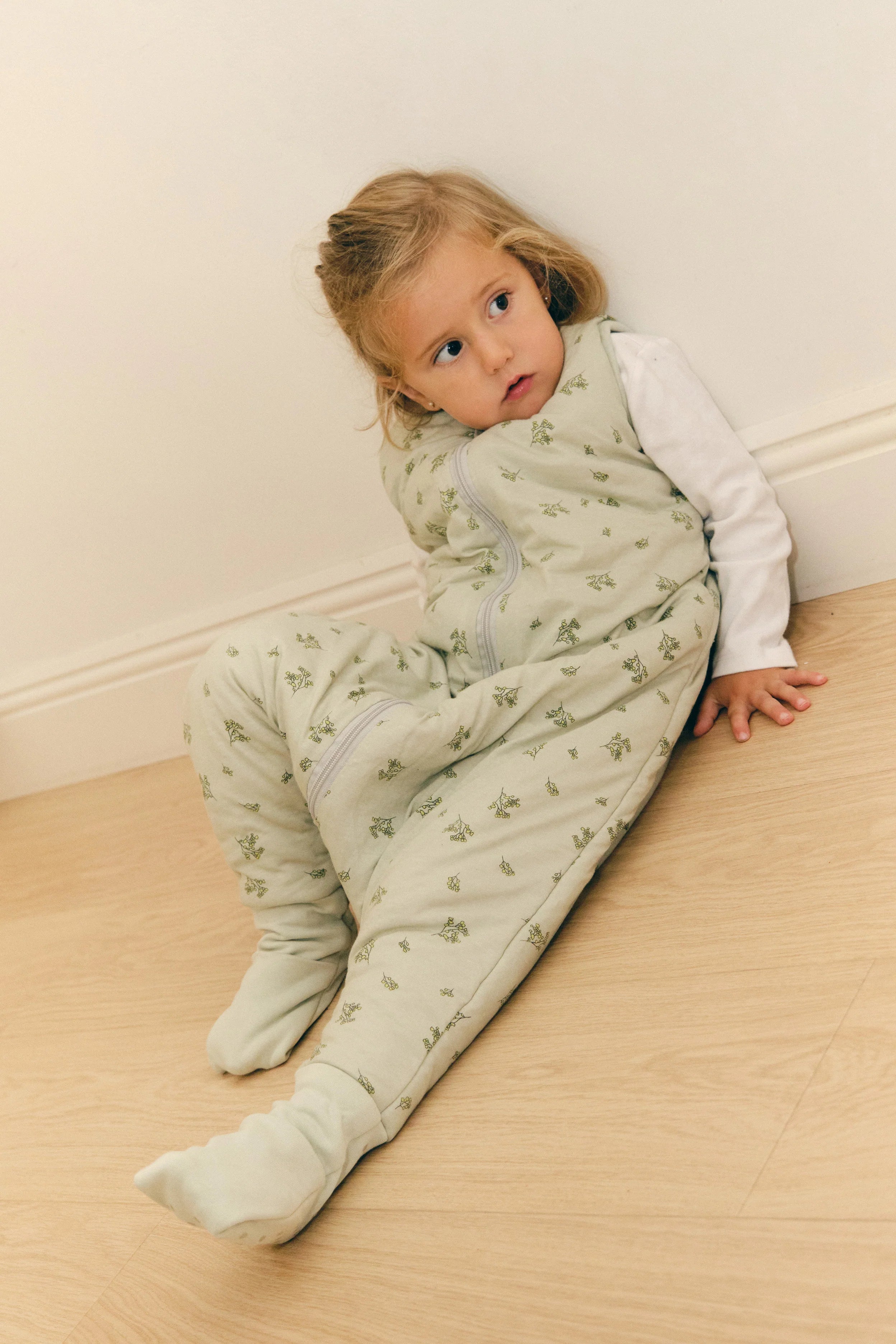 Menina sentada no chão, encostada à parede, com saco de dormir MolisandCo TOG 2.5 com pés Botany Green e meias antiderrapantes.