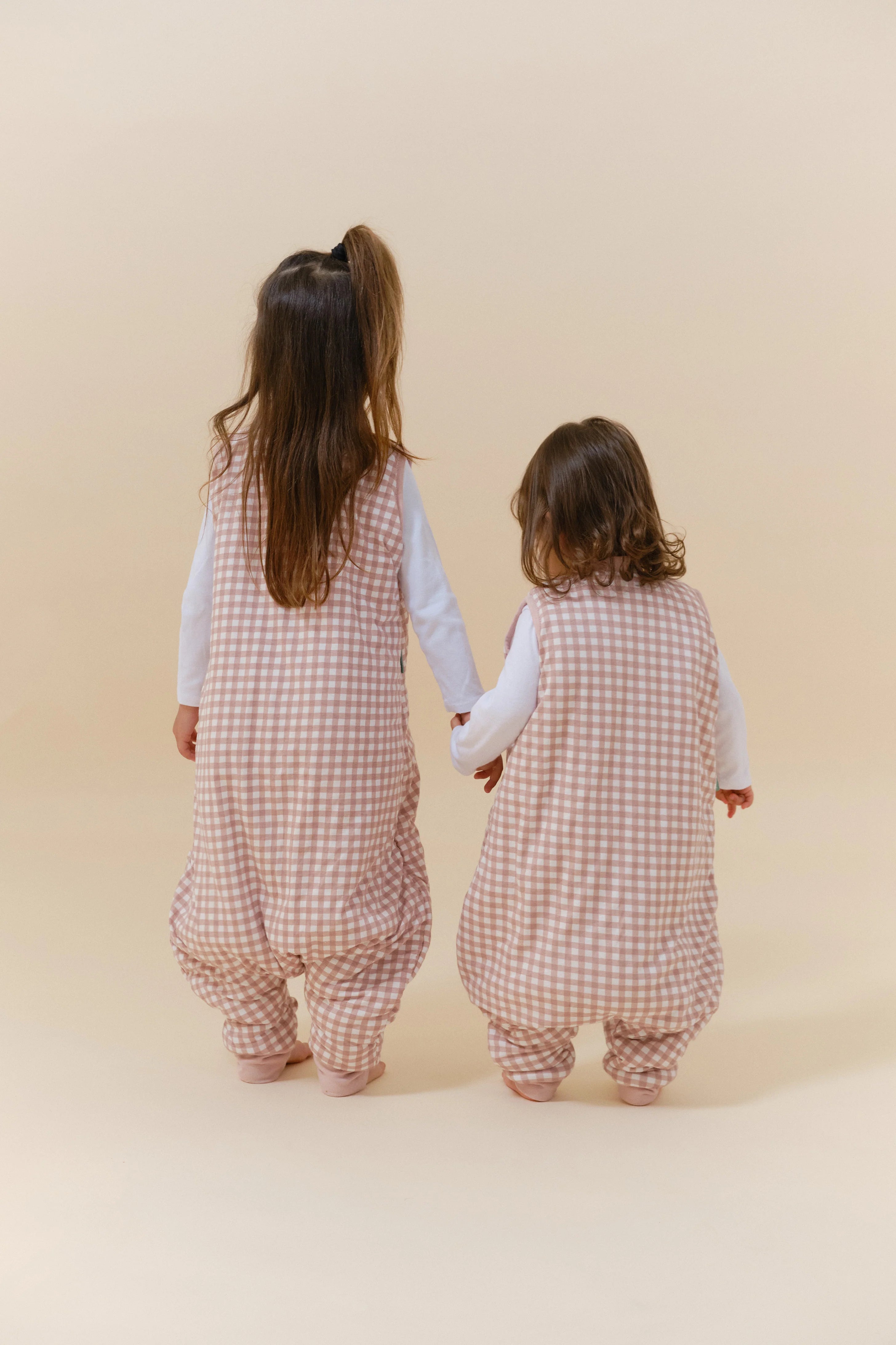 Duas crianças de costas, de mãos dadas, com sacos de dormir MolisandCo TOG 2.5 com pés rosa xadrez.
