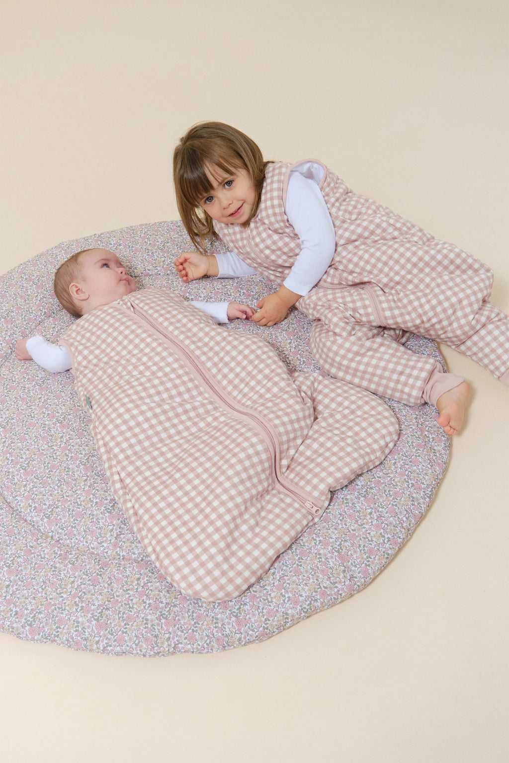 Duas crianças, bebé e criança mais velha, deitadas em tapete, com sacos de dormir MolisandCo TOG 2.5 com pés rosa xadrez.