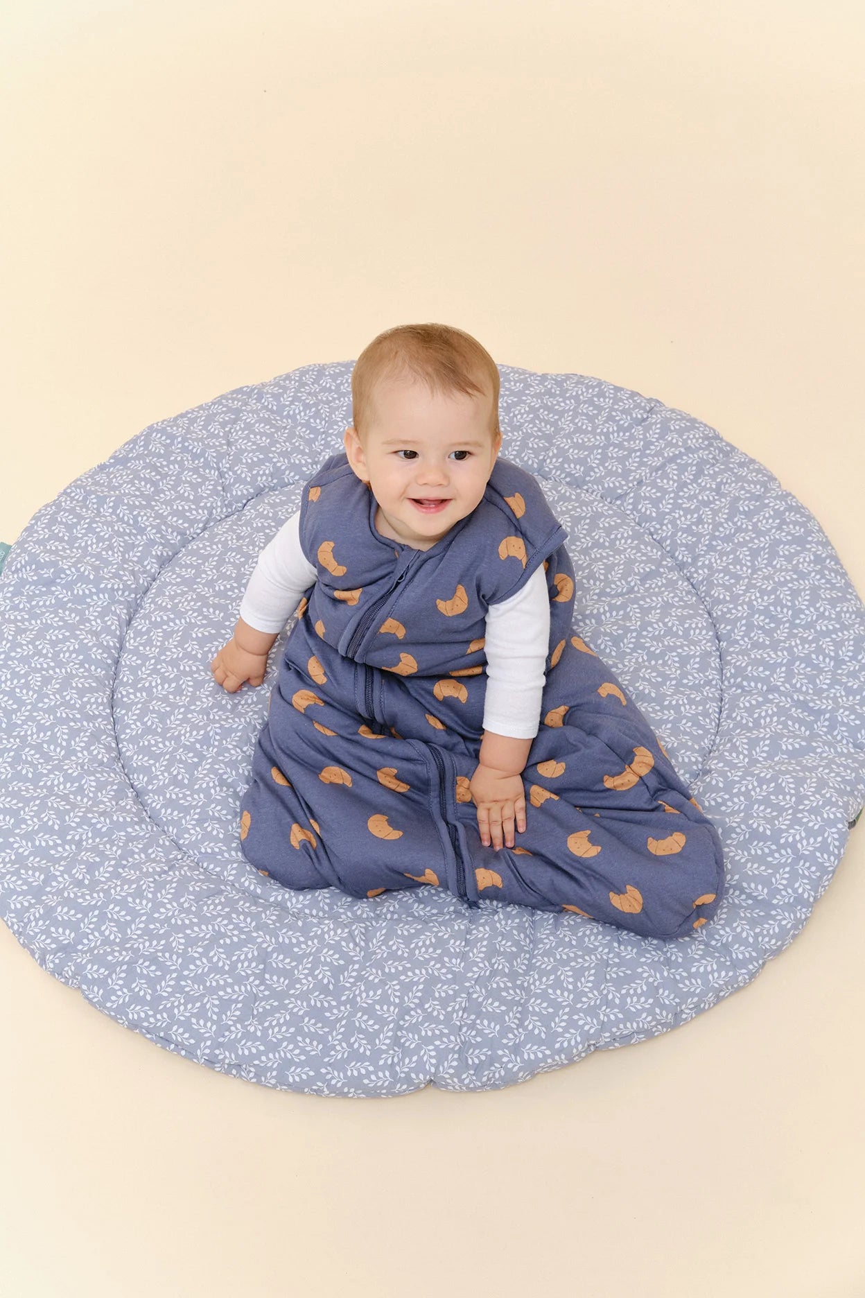 Bebé sorridente em saco de dormir MolisandCo TOG 2.5 azul com padrão croissants, sentado num tapete redondo.