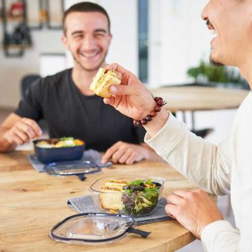 Dois homens a partilhar uma refeição descontraída, um com a marmita Monbento cheia, o outro com uma salada na lancheira.