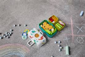 Lancheira Monbento Tresor Dinossauros aberta, com snacks coloridos, disposta num chão de cimento com giz e berlindes, simulando um ambiente de brincadeira.